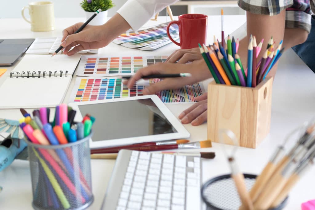 Graphic design workspace with colour swatches, pencils, markers, tablet, keyboard and creative tools used during the design process.