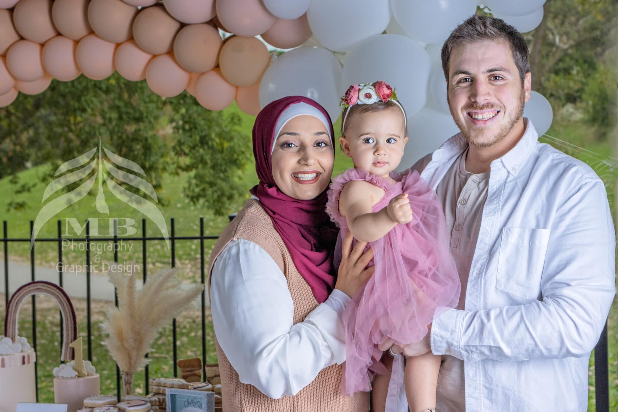 Photography Portfolio 53 Mariam Cheaitou Bazzi Photography Portfolio in Windsor Mother and father holding their baby girl in a pink dress beneath a pastel balloon display during a first-birthday family photography session in Windsor, Ontario.