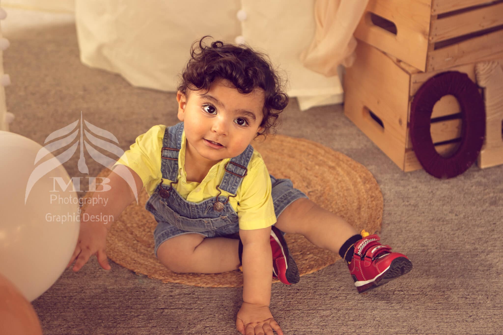 Photography Portfolio 32 Mariam Cheaitou Bazzi Photography Portfolio in Windsor One year old boy in overalls sitting on jute rug reaching toward balloon, first birthday photography by Windsor Ontario photographer