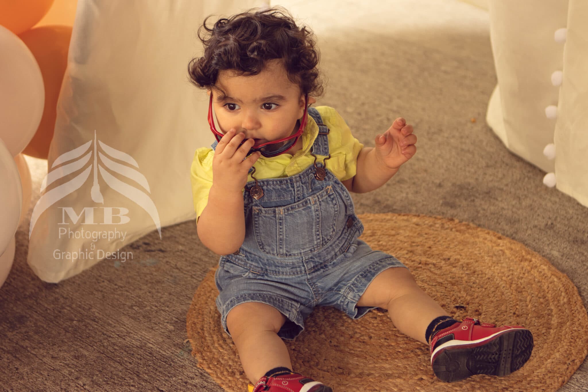 Photography Portfolio 31 Mariam Cheaitou Bazzi Photography Portfolio in Windsor One year old boy in overalls playfully chewing on sunglasses while sitting on jute rug, first birthday photography by Windsor Ontario photographer