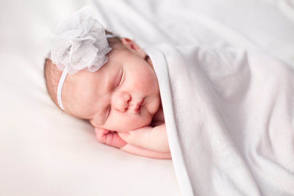 Newborn photography in Windsor Ontario. Newborn Baby Girl Sleeping Peacefully Under Blanket, Wearing Flower Headband.