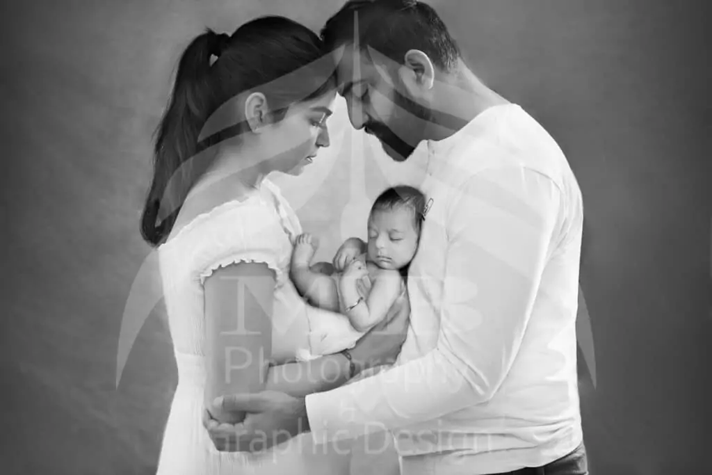 Black and white newborn photography session in Windsor, Ontario featuring a mother and father holding their sleeping baby in a soft, intimate studio portrait by MB Graphic & Photography Design.