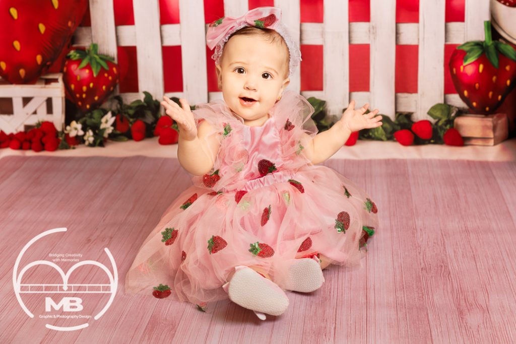 Milestone baby plan photography session in Windsor, Ontario by MB Graphic & Photography Design, strawberry-themed  featuring a smiling baby girl in a pink strawberry dress sitting on a pink floor with red strawberry backdrop.
