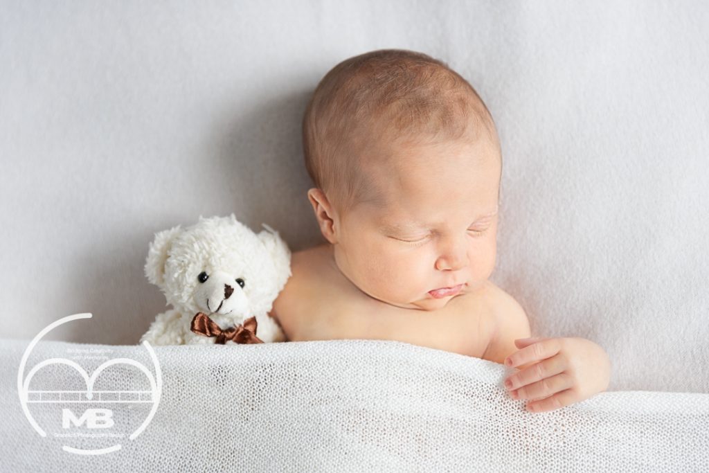 Newborn baby photography session in Windsor, Ontario by MB Graphic & Photography Design, featuring a peaceful sleeping baby wrapped in a soft white blanket beside a small teddy bear on a clean neutral backdrop.
