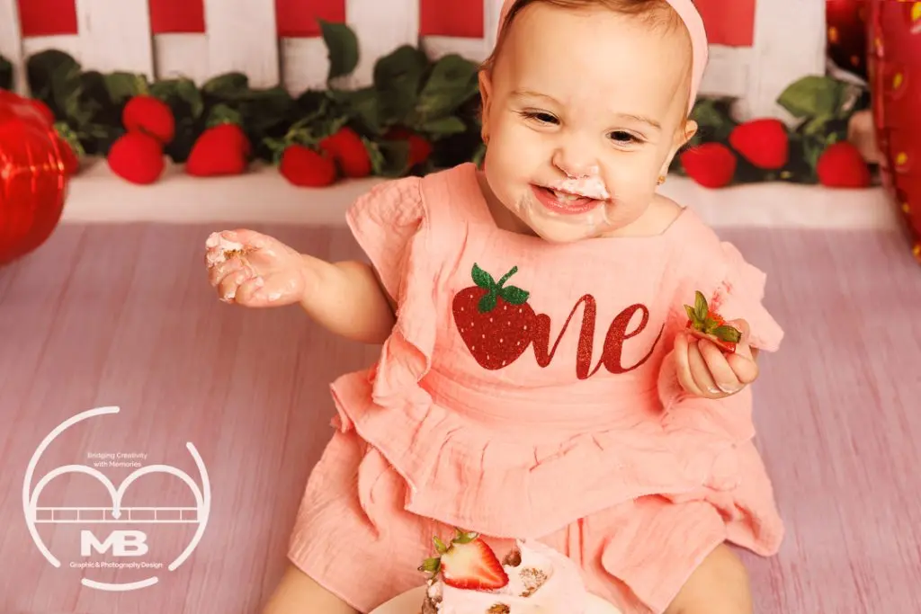 First birthday cake smash photography session in Windsor, Ontario featuring a smiling baby girl in a strawberry-themed outfit enjoying cake with a red and pink backdrop by MB Graphic & Photography Design.
