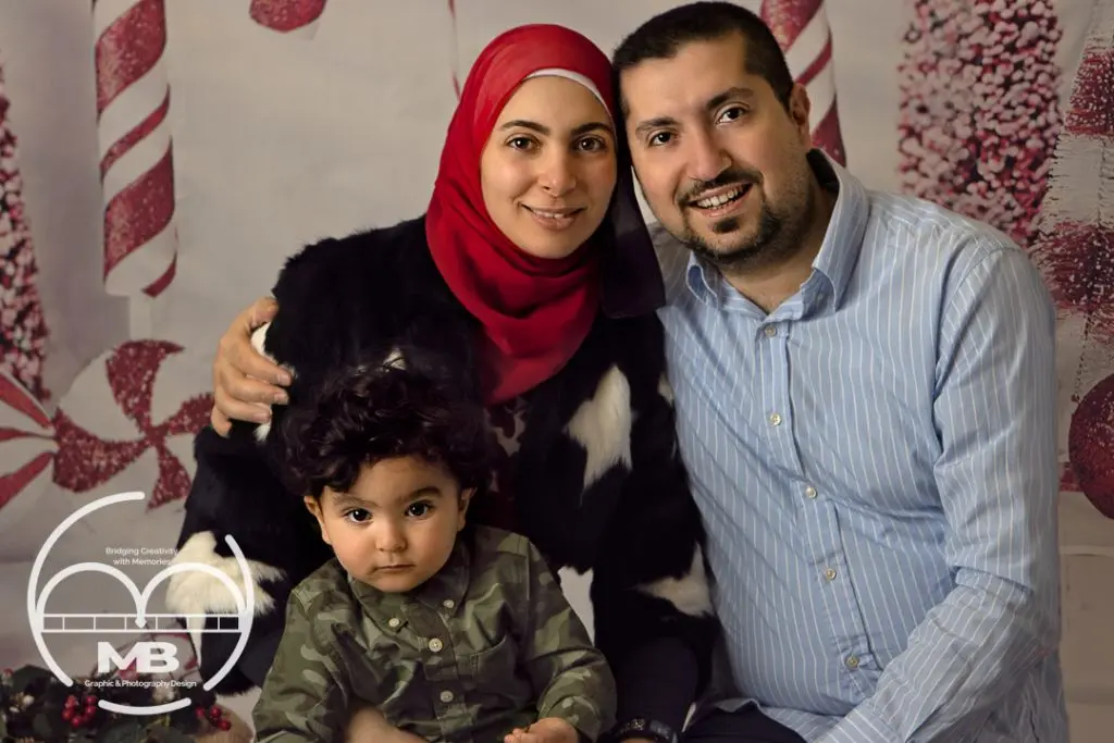 Family photography session in Windsor, Ontario featuring a smiling mother in a red hijab, father, and young child posed together in a festive holiday studio setting by MB Graphic & Photography Design.